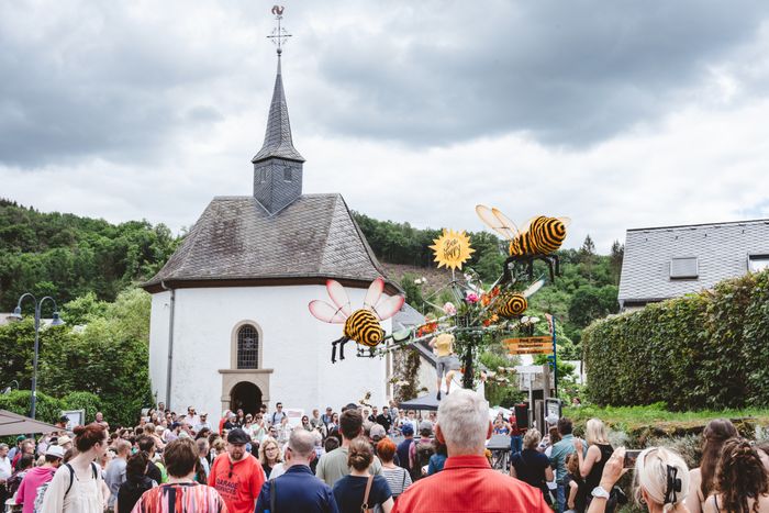 Ein belebter Platz mit Kirchturm im Ortszentrum von Lellingen während des Open Air Konstfestivals. Menschen schlendern zwischen Skulpturen und Kunstwerken.